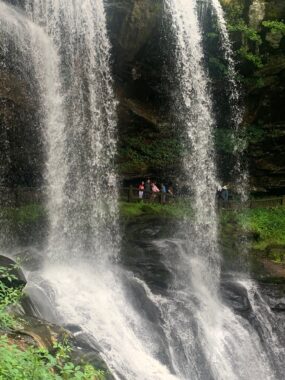The photo shows a large waterfall cascading over rocks, with the moisture allowing for moss and plants to thrive. Behind the falls is a narrow walkway. We can see three people posing for a photo between streams of water, but they appear small in the distance.